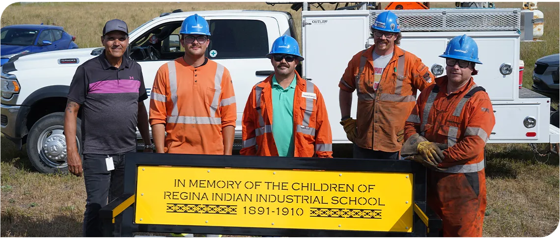 Five men stand behind a yellow bench. Four of the men wear orange work shirts and blue hard hats.