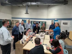 A group of people stand around a table while touring a facility. At the table sit workers from Cosmo industries, taking apart SaskEnergy's AMI modules.