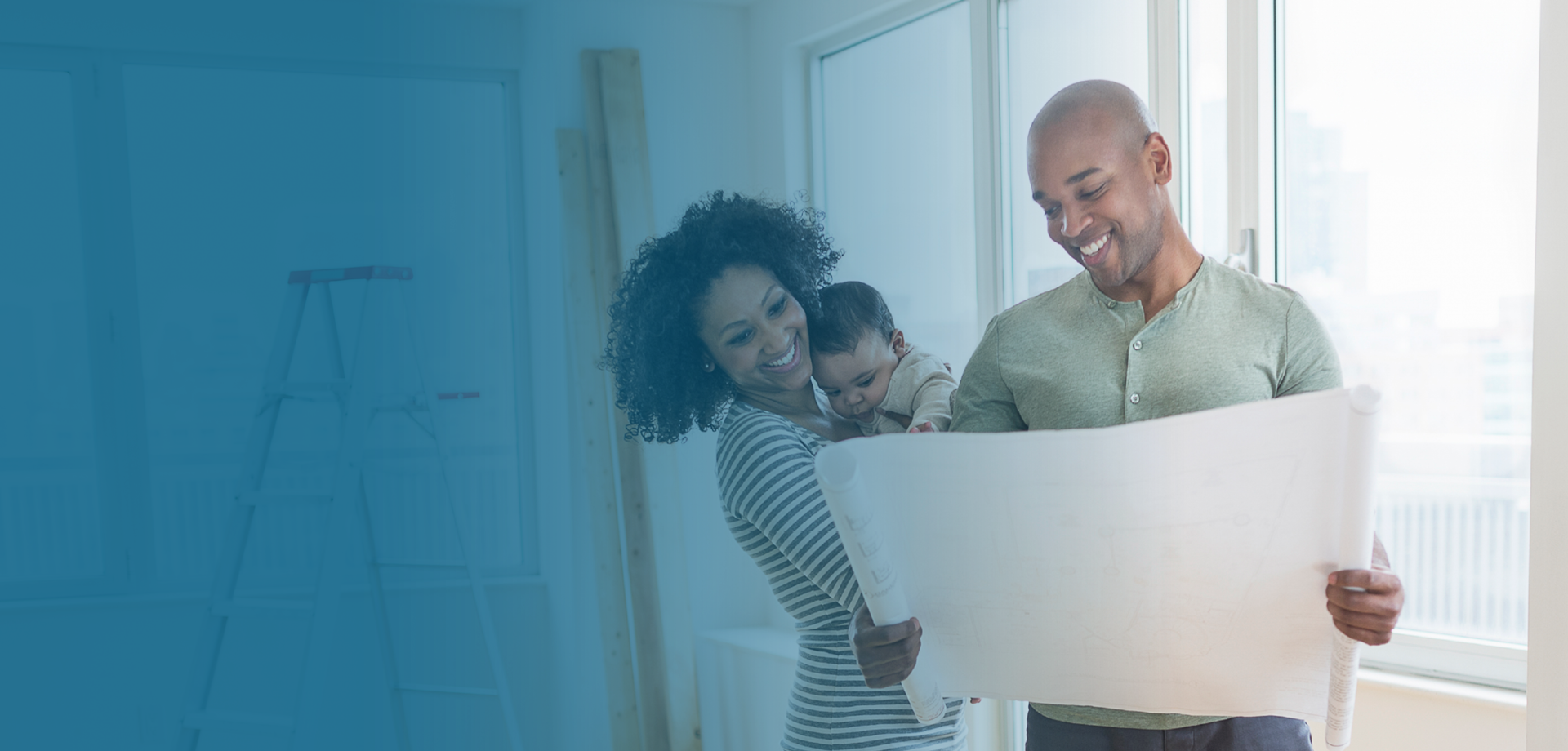 couple standing in their home being built looking at a floor plan and smiling with their baby