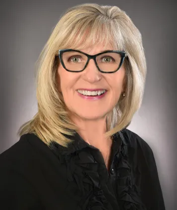 Susan Barber smiles - she wears a black top and has shoulder-length blonde hair and glasses.