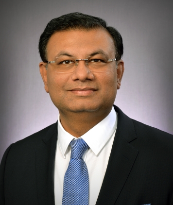 A man with glasses and short black hair, in a black suit jacket, white shirt and blue tie. 